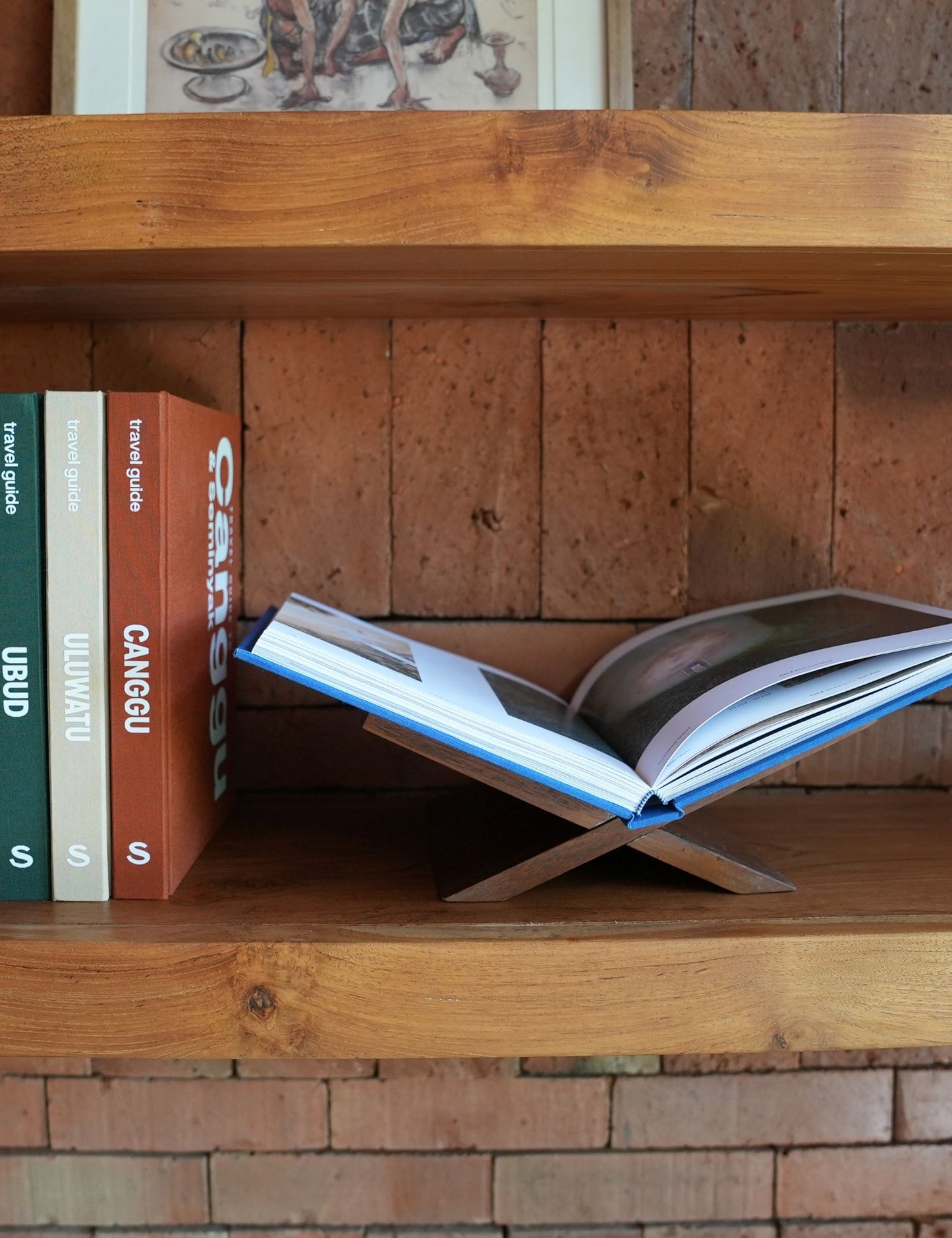 Open book on a wooden shelf with other books stacked behind, against a brick wall.